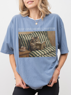 Woman wearing a blue jean t shirt with just one more chapter text and vintage style illustration of a cat reading a book on a striped couch with a teacup beside it