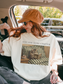 Woman sitting in a car wearing an ivory t shirt with just one more chapter text and a cozy cat reading a book illustration on a striped couch