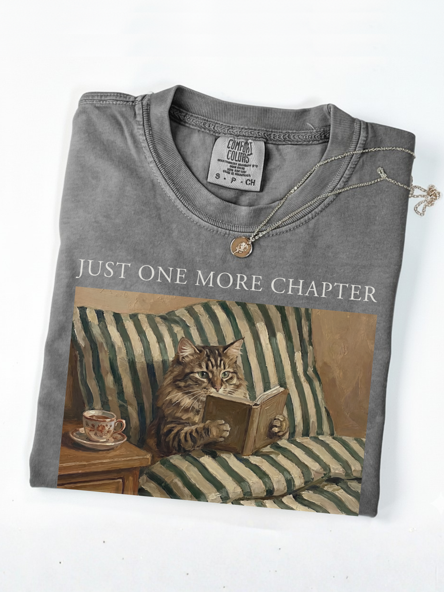 Folded pepper t shirt with just one more chapter text and vintage painting of a cat reading a book on a couch with a teacup styled with a gold necklace