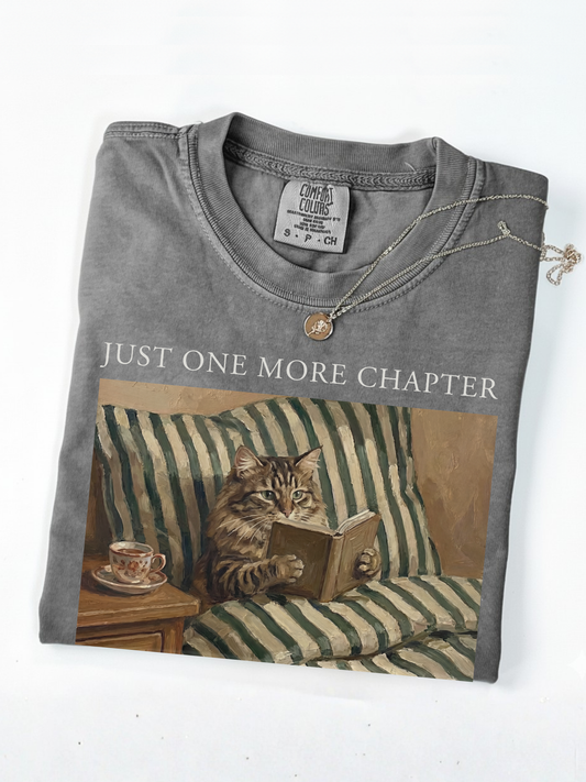 Folded pepper t shirt with just one more chapter text and vintage painting of a cat reading a book on a couch with a teacup styled with a gold necklace