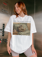 Woman standing in an elevator wearing a white t shirt with just one more chapter text and a vintage illustration of a cat reading a book on a couch
