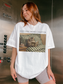 Woman standing in an elevator wearing a white t shirt with just one more chapter text and a vintage illustration of a cat reading a book on a couch