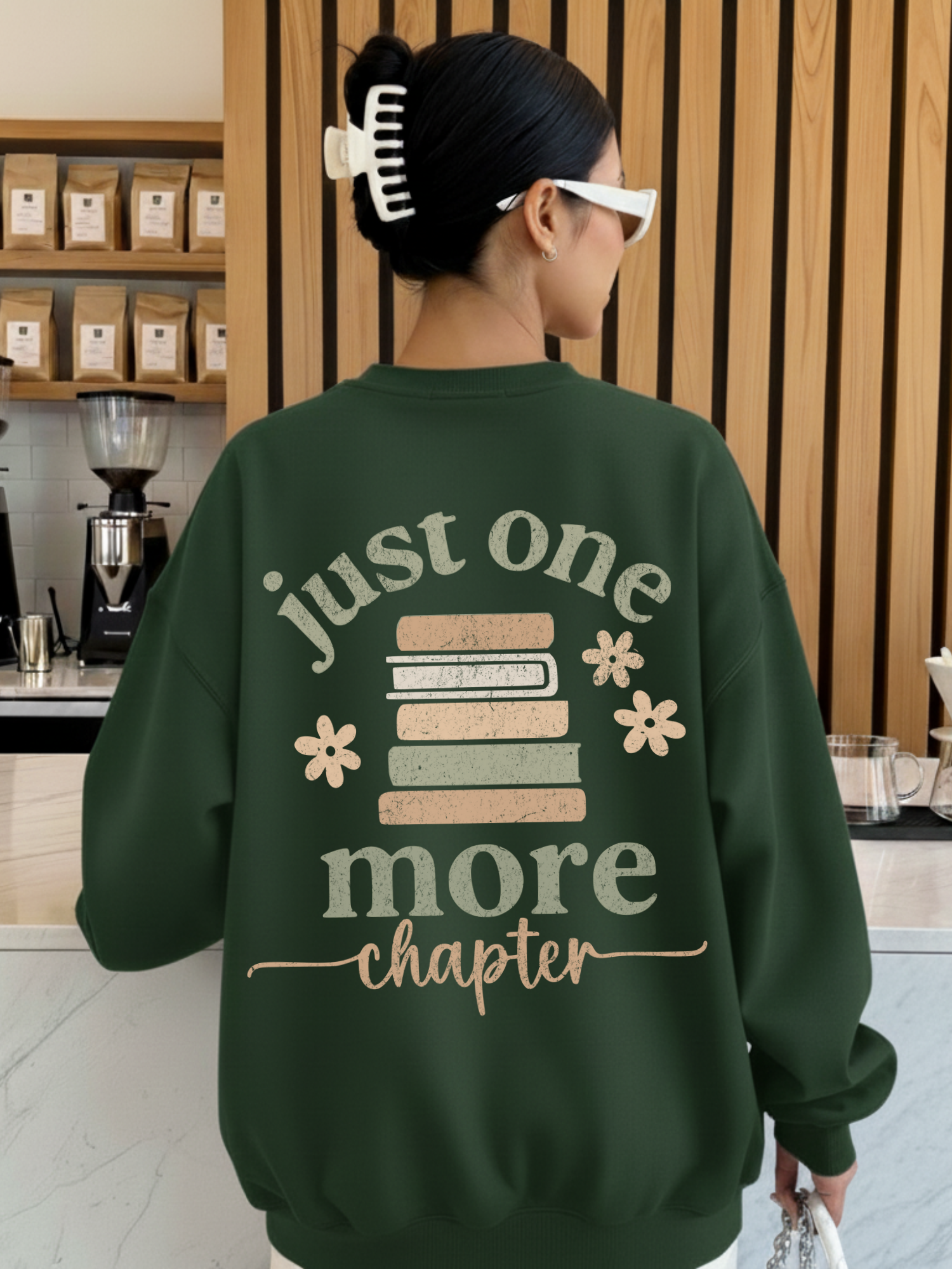 Woman wearing a forest green sweatshirt featuring a large back design with stacked pastel books, floral accents, and the phrase just one more chapter in textured script. Styled in a cozy coffee shop with wooden walls and espresso gear in the background.