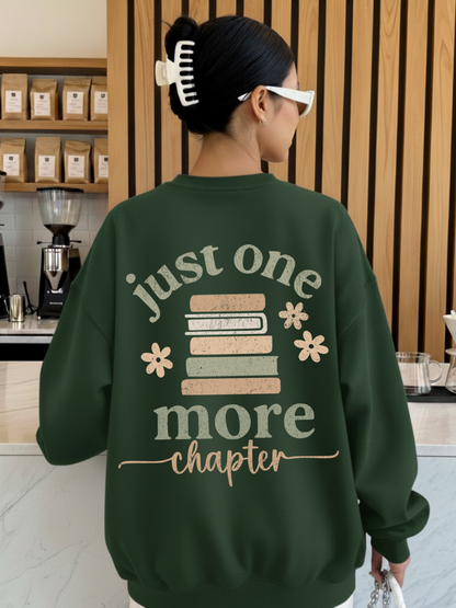 Woman wearing a forest green sweatshirt featuring a large back design with stacked pastel books, floral accents, and the phrase just one more chapter in textured script. Styled in a cozy coffee shop with wooden walls and espresso gear in the background.
