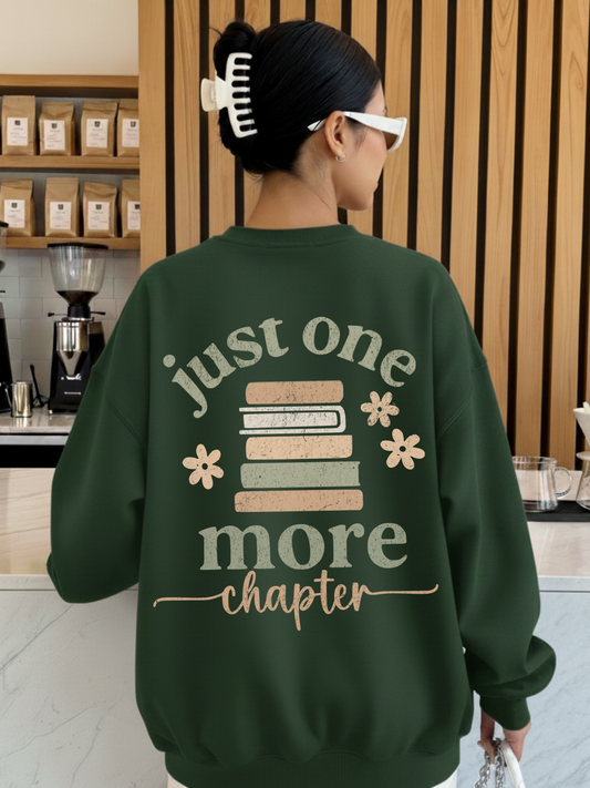 Woman wearing a forest green sweatshirt featuring a large back design with stacked pastel books, floral accents, and the phrase just one more chapter in textured script. Styled in a cozy coffee shop with wooden walls and espresso gear in the background.