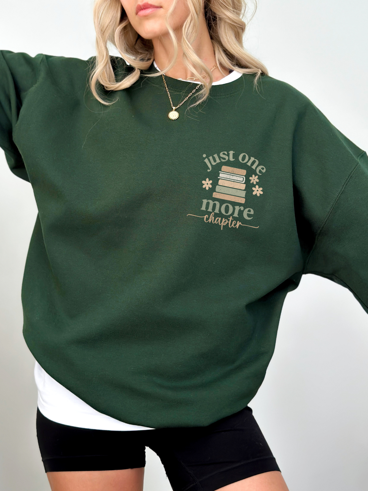 Woman wearing a forest green sweatshirt styled with black biker shorts. The sweatshirt features a cozy front chest graphic with a beige and blush book stack, daisies, and the text just one more chapter in cursive and serif.