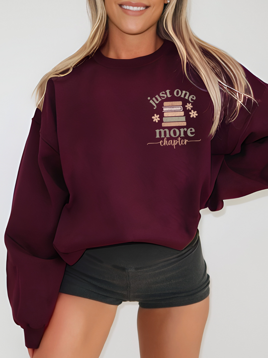 Woman wearing a maroon sweatshirt with a minimalist front chest graphic showing a pastel book stack, daisy accents, and the phrase just one more chapter in serif and script fonts. Styled with black shorts in a studio setting.
