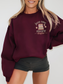 Woman wearing a maroon sweatshirt with a minimalist front chest graphic showing a pastel book stack, daisy accents, and the phrase just one more chapter in serif and script fonts. Styled with black shorts in a studio setting.
