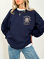 Woman in a navy crewneck sweatshirt featuring a front left chest design with stacked vintage-style books, soft florals, and just one more chapter in warm neutral lettering. Styled with light blue jeans and white tee underneath.