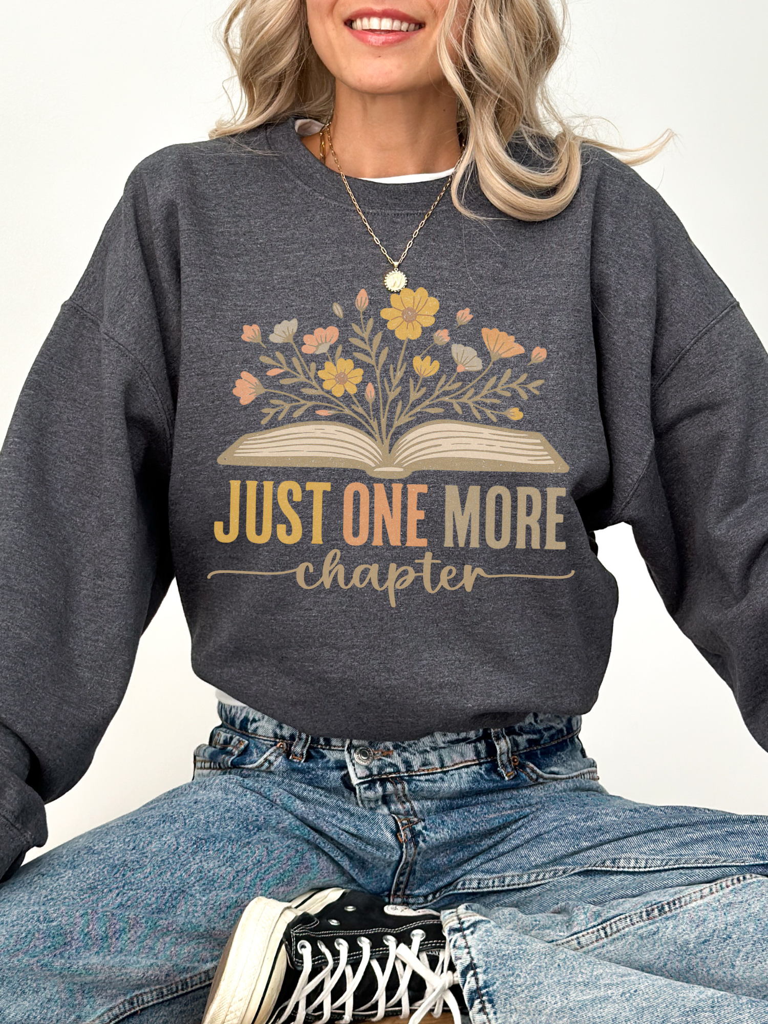 Woman wearing dark heather Gildan sweatshirt that features an open book with pastel floral bouquet and the phrase just one more chapter in warm tones, seated cross-legged in light blue jeans and gold pendant necklace