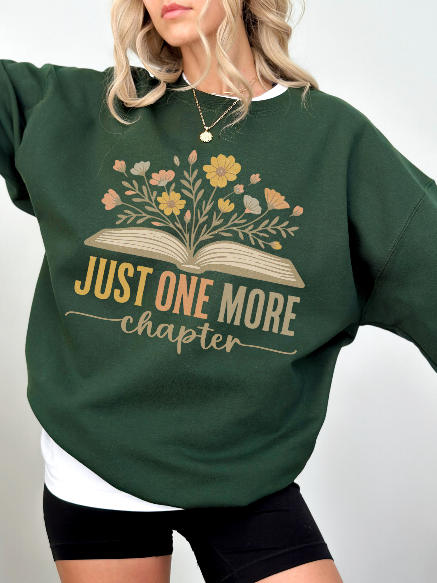 Woman in forest green cozy sweatshirt featuring vintage-style floral book graphic and just one more chapter quote, layered over a white shirt and styled with black shorts, standing with hand on hip