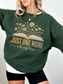 Woman in forest green cozy sweatshirt featuring vintage-style floral book graphic and just one more chapter quote, layered over a white shirt and styled with black shorts, standing with hand on hip