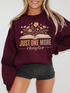 Woman wearing maroon Gildan sweatshirt with floral book design that says just one more chapter in retro pastel text, standing against a white wall with hands at sides, styled with black shorts and blonde hair