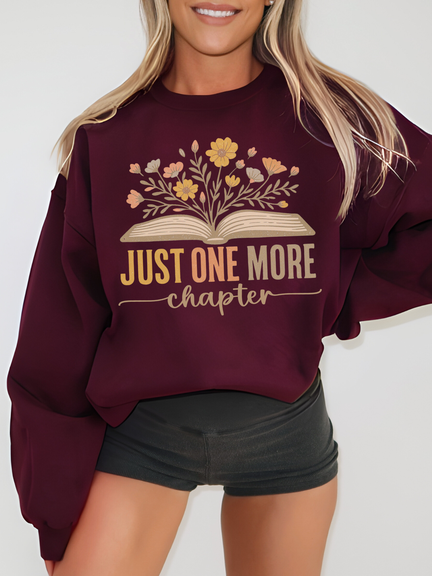 Woman wearing maroon Gildan sweatshirt with floral book design that says just one more chapter in retro pastel text, standing against a white wall with hands at sides, styled with black shorts and blonde hair