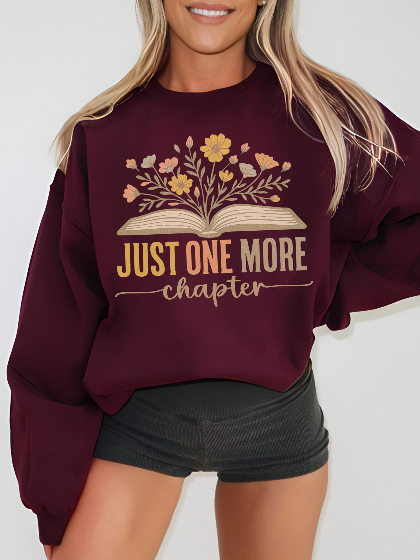 Woman wearing maroon Gildan sweatshirt with floral book design that says just one more chapter in retro pastel text, standing against a white wall with hands at sides, styled with black shorts and blonde hair