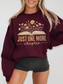 Woman wearing maroon Gildan sweatshirt with floral book design that says just one more chapter in retro pastel text, standing against a white wall with hands at sides, styled with black shorts and blonde hair