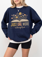 Woman in navy blue sweatshirt with floral book design and text just one more chapter, styled with black biker shorts and layered white tee, standing casually with relaxed posture and light curled hair