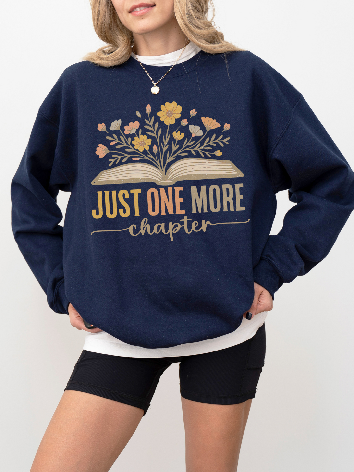 Woman in navy blue sweatshirt with floral book design and text just one more chapter, styled with black biker shorts and layered white tee, standing casually with relaxed posture and light curled hair