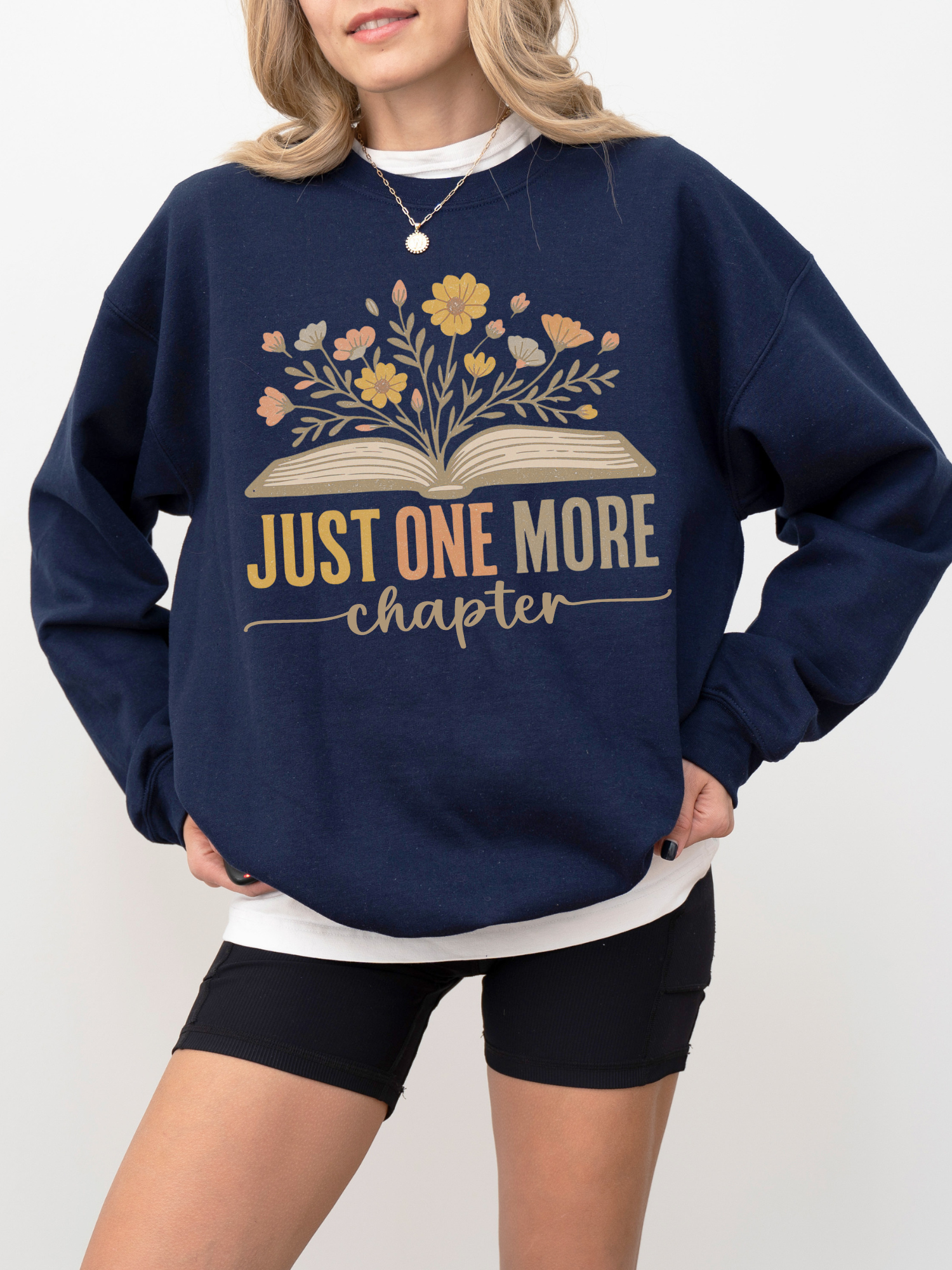 Woman in navy blue sweatshirt with floral book design and text just one more chapter, styled with black biker shorts and layered white tee, standing casually with relaxed posture and light curled hair