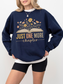 Woman in navy blue sweatshirt with floral book design and text just one more chapter, styled with black biker shorts and layered white tee, standing casually with relaxed posture and light curled hair