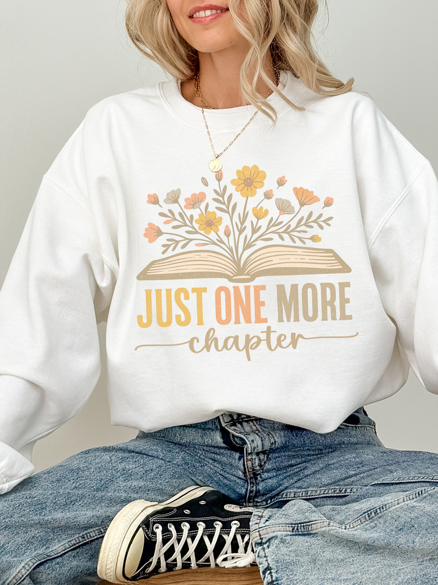 Woman in crisp white Gildan sweatshirt showing off pastel floral book artwork and the text just one more chapter in soft yellow and peach hues, seated on wooden stool in light-washed jeans and black sneakers