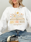 Woman in crisp white Gildan sweatshirt showing off pastel floral book artwork and the text just one more chapter in soft yellow and peach hues, seated on wooden stool in light-washed jeans and black sneakers