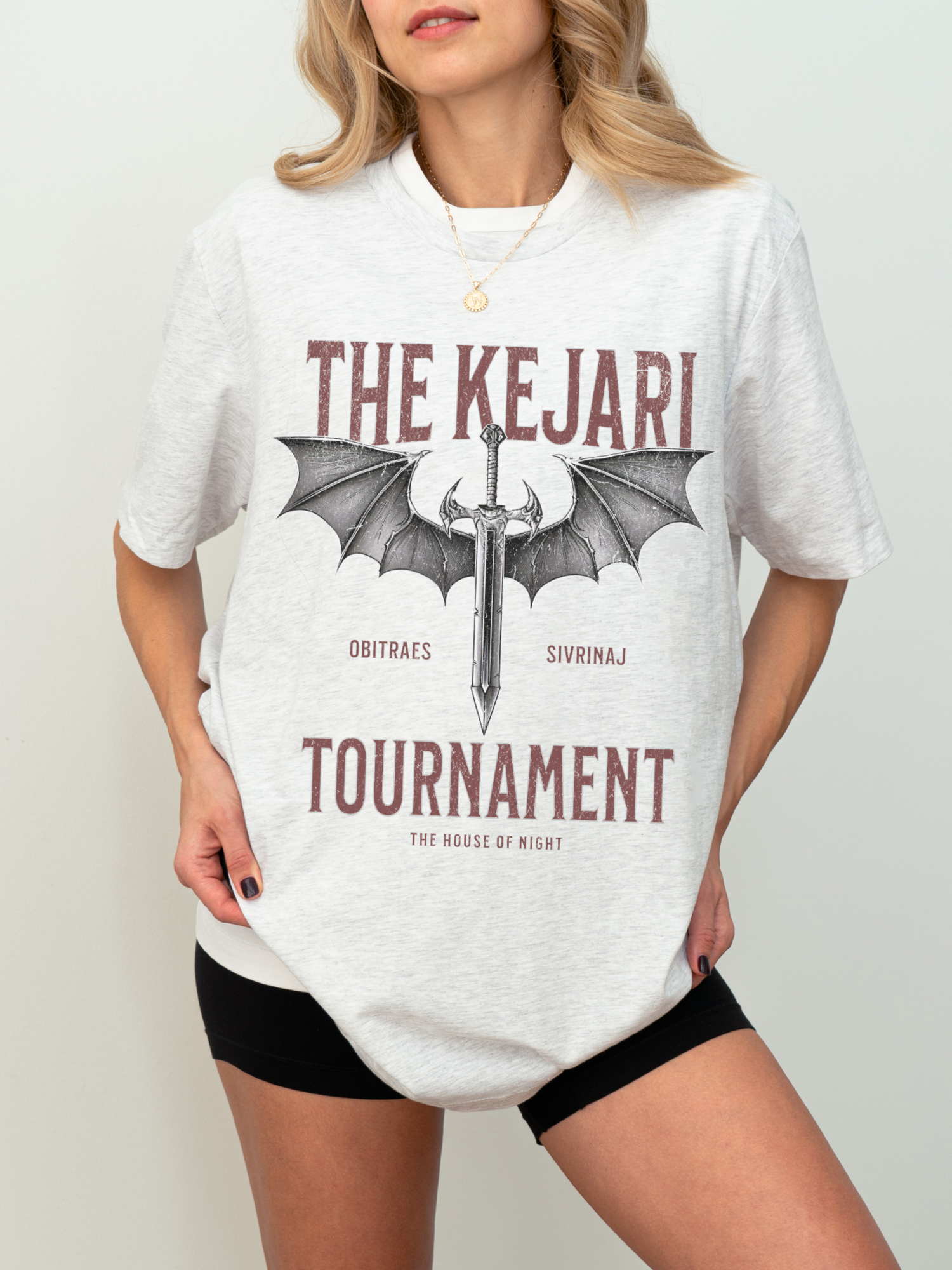 Woman wearing ash grey Kejari Tournament t-shirt styled for fantasy book fans, featuring sword and wing graphic design and subtle red-brown vintage typography.