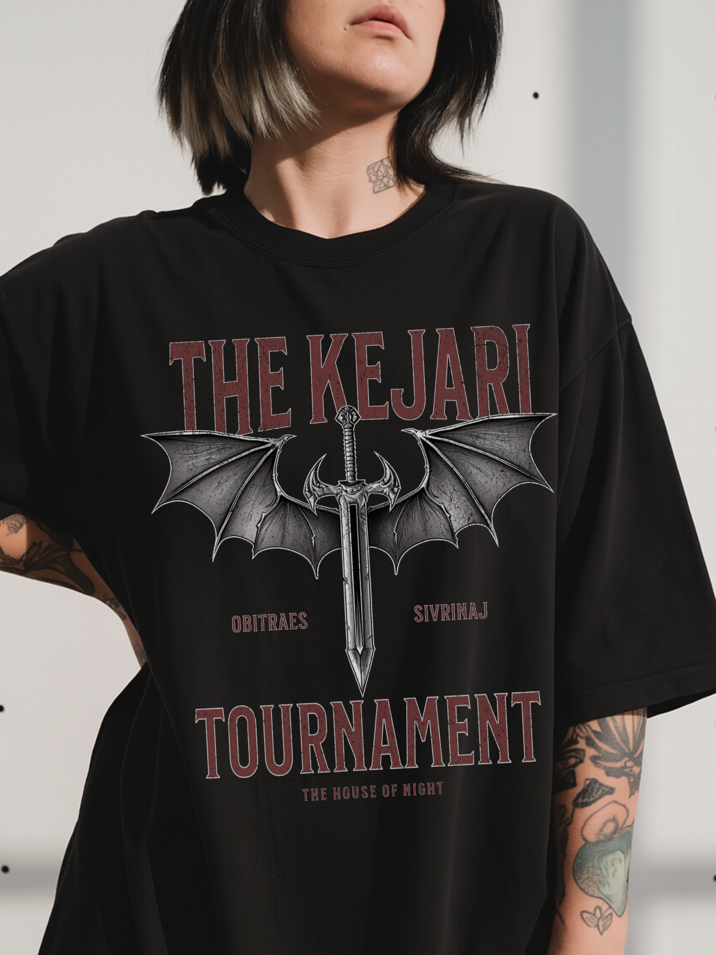 Close-up of a woman wearing a black Kejari Tournament fantasy graphic tee featuring distressed text and dragon-winged sword artwork inspired by the House of Night series by Carissa Broadbent. Bold red and gray typography adds a vintage battle aesthetic.