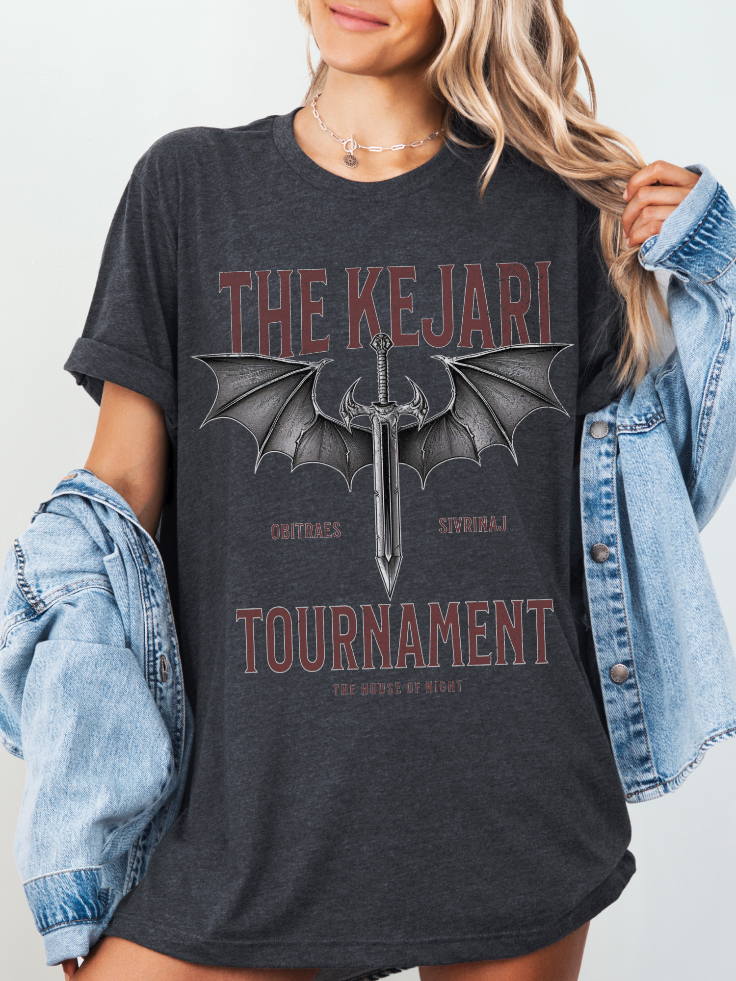 Dark heather grey Kejari Tournament t-shirt worn with denim jacket, featuring gothic-style dragon wings and fantasy sword illustration for romantasy fans.