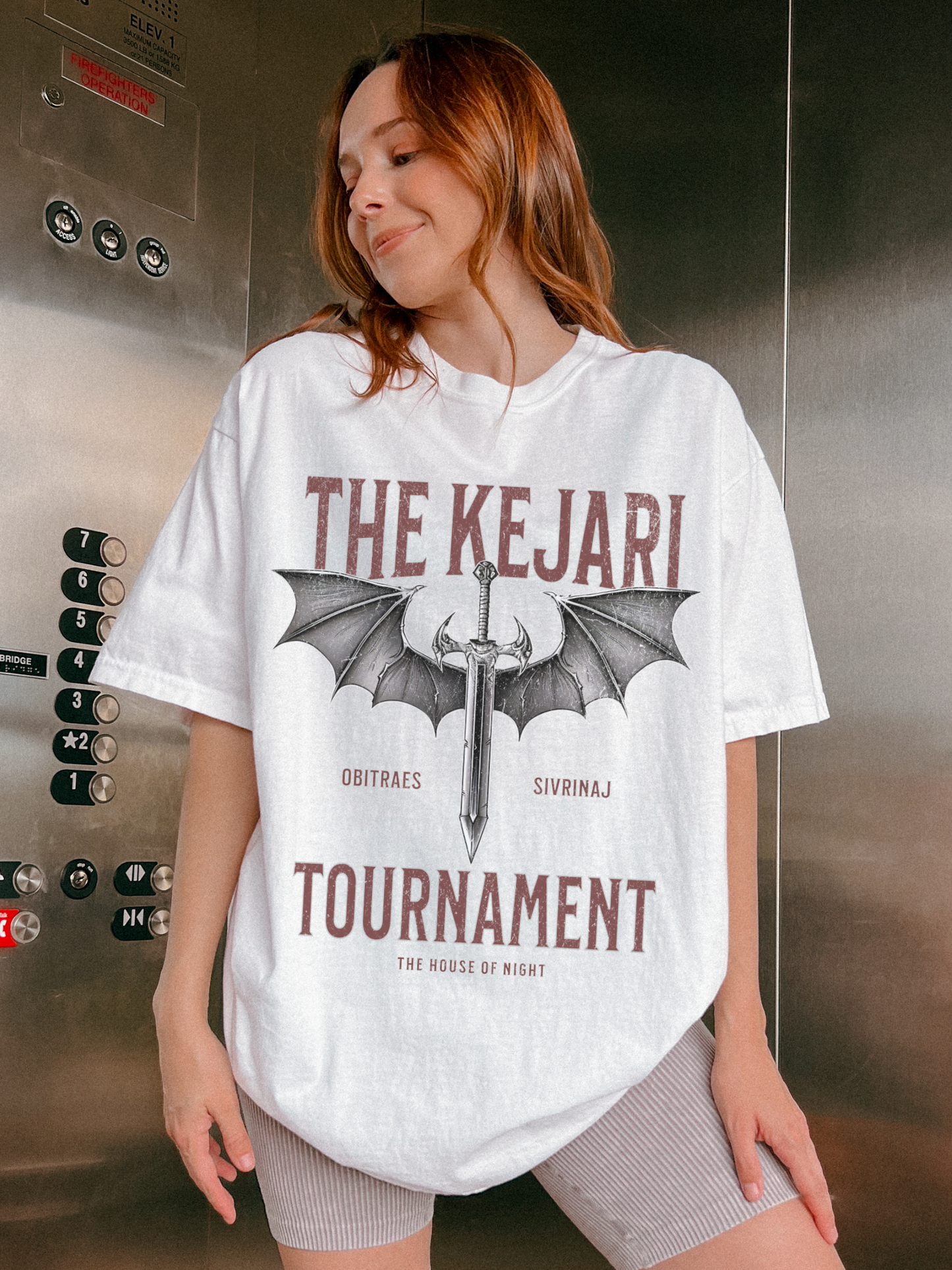 Woman wearing a white oversized fantasy book tee in an elevator, featuring distressed Kejari Tournament artwork with dragon wings and a blade, styled for bookish streetwear vibes.