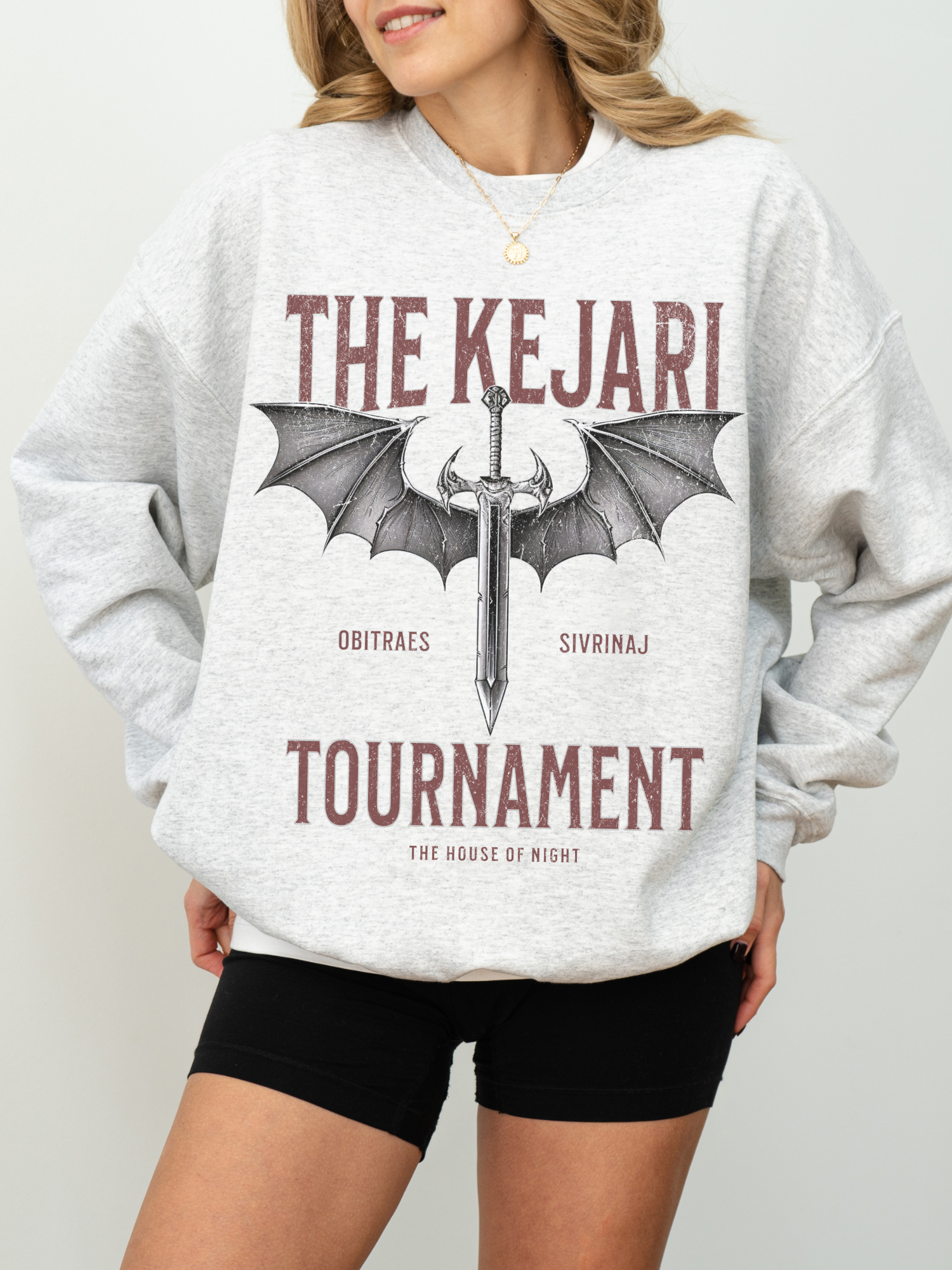 Ash Grey Kejari Tournament fantasy sweatshirt shown on a woman, bold maroon text and gothic sword graphic visible, worn with black shorts