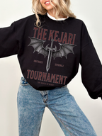 Woman posing in black Kejari Tournament sweatshirt with fantasy sword and wings print, paired with light blue jeans and gold necklace