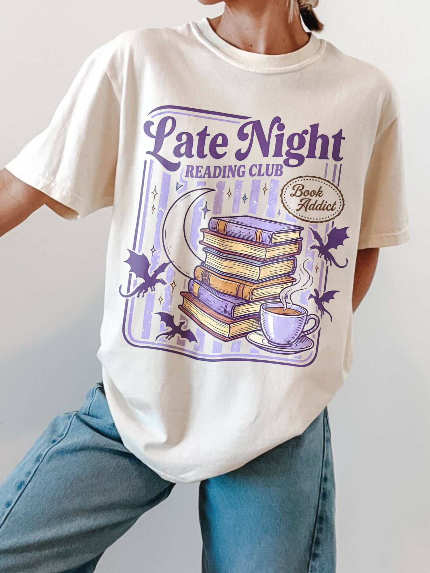 Woman wearing oversized Comfort Colors late night reading club shirt featuring retro stacked books crescent moon bats and coffee cup graphic in purple tones styled with light wash jeans against neutral background