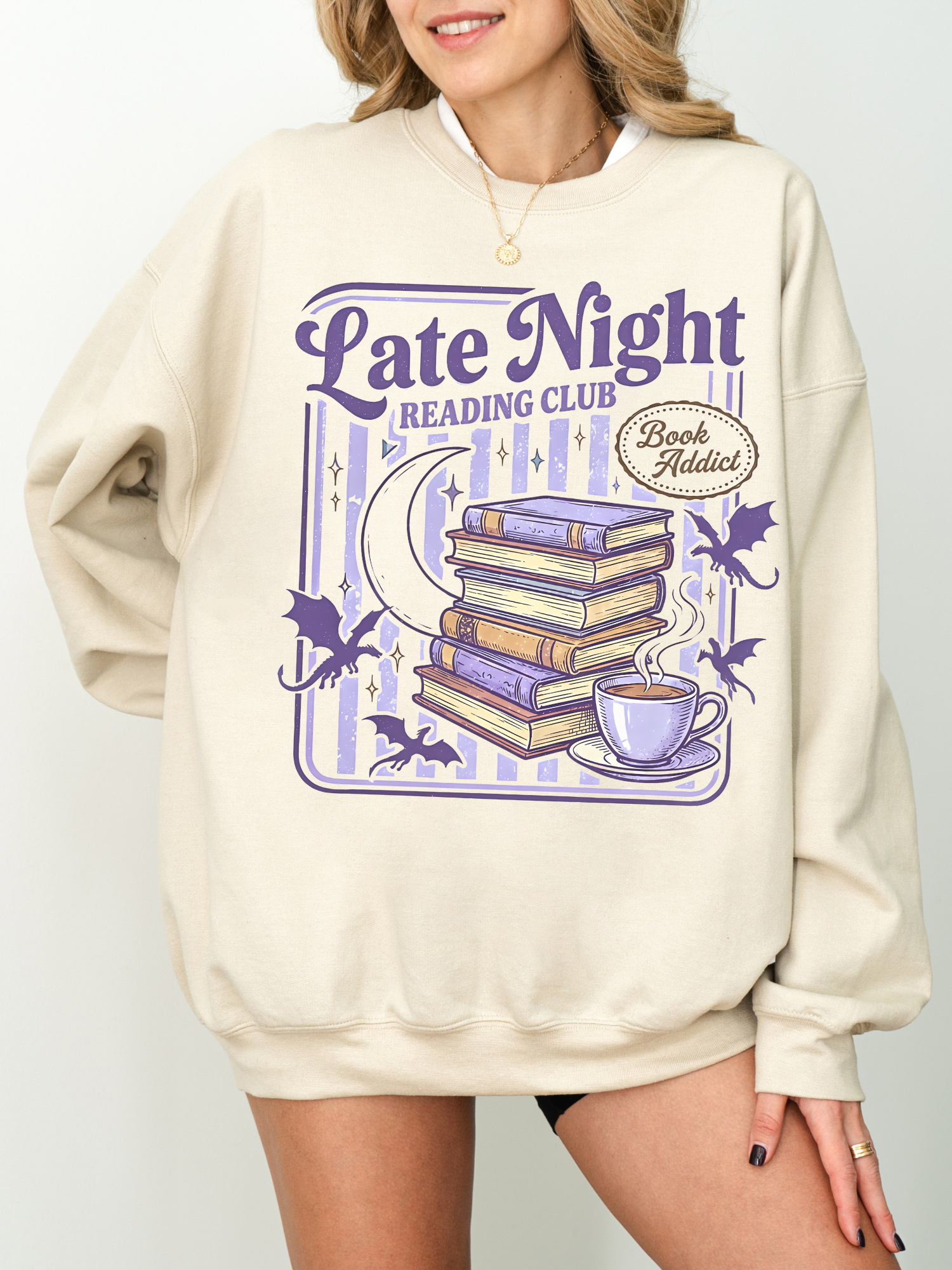 Woman wearing oversized beige late night reading club sweatshirt featuring retro stacked books crescent moon bats and coffee cup graphic in purple tones styled with minimal jewelry