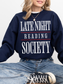 woman sitting in a navy sweatshirt with bold white and red text that says late night reading society