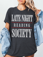 woman wearing a dark heather grey tee that says late night reading society in bold white font with red lines and denim jacket