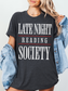 woman wearing a dark heather grey tee that says late night reading society in bold white font with red lines and denim jacket