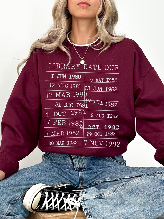 woman sitting cross-legged wearing maroon library due date sweatshirt with white print
