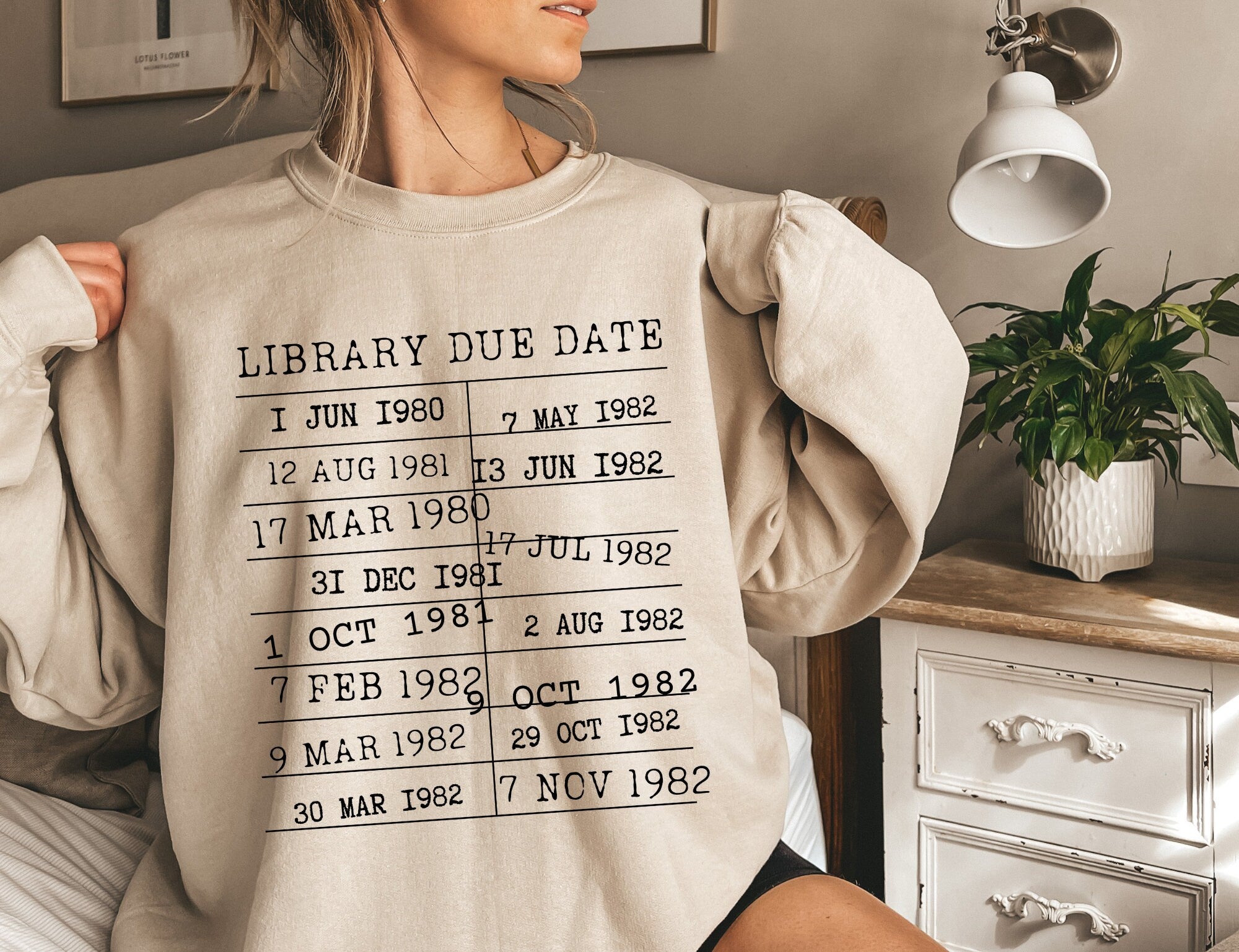 woman lounging in sand-colored library due date sweatshirt with vintage book stamp design