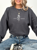 Front view of woman in dark heather grey sweatshirt with small chest graphic of snake curled around rose and the words Little Serpent, fantasy book merch inspired by The Serpent and the Wings of Night