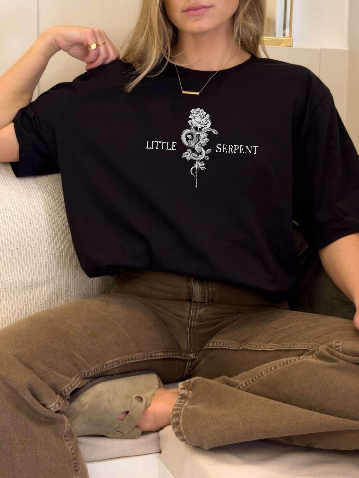 Casual lifestyle photo of a black t-shirt featuring a central rose and serpent artwork with Little Serpent text on each side, worn by a model lounging on a couch in relaxed fantasy reader style.