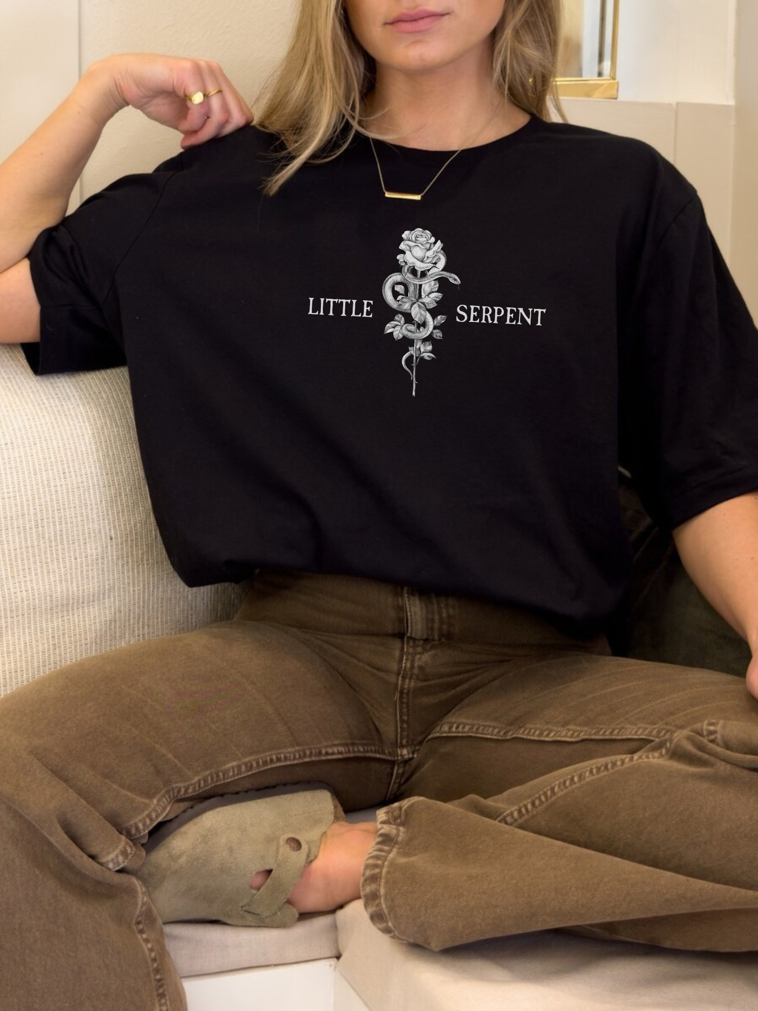 Casual lifestyle photo of a black t-shirt featuring a central rose and serpent artwork with Little Serpent text on each side, worn by a model lounging on a couch in relaxed fantasy reader style.