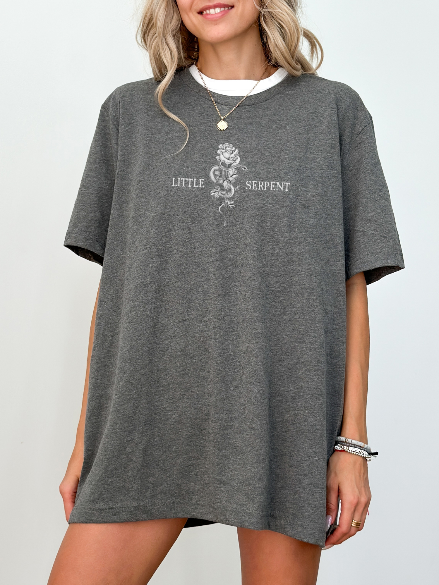 Model wearing a dark heather grey t-shirt with a minimalist fantasy design showing a snake and rose illustration between the words Little Serpent, officially licensed The Serpent and the Wings of Night apparel.