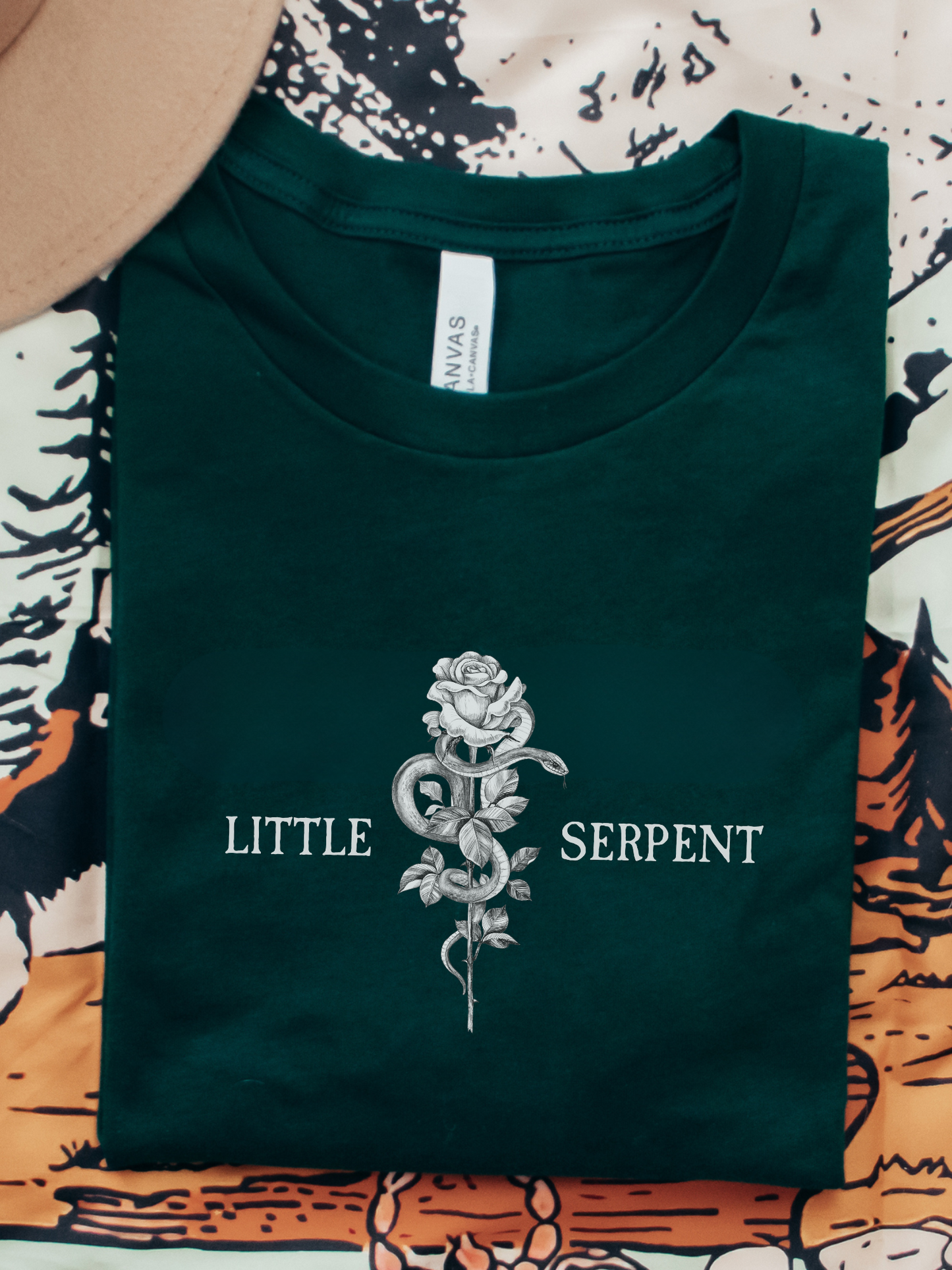 Flatlay of a forest green Bella Canvas t-shirt featuring an intricate rose and snake design from Crowns of Nyaxia merchandise, laid out beside a neutral-toned hat and fantasy book-themed blanket.