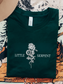 Flatlay of a forest green Bella Canvas t-shirt featuring an intricate rose and snake design from Crowns of Nyaxia merchandise, laid out beside a neutral-toned hat and fantasy book-themed blanket.