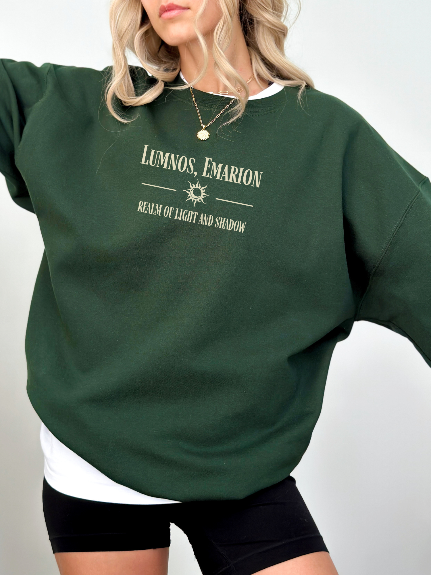 Woman modeling a forest green Gildan sweatshirt with “Lumnos, Emarion” quote print, styled with black biker shorts and white layering.