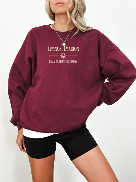 Woman wearing a maroon fantasy-themed Gildan sweatshirt reading “Lumnos, Emarion – Realm of Light and Shadow,” with a relaxed, cozy aesthetic.