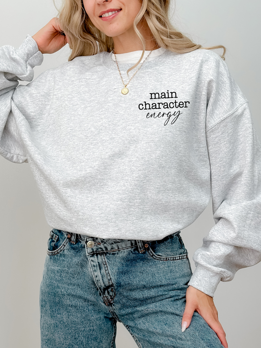 woman in ash grey sweatshirt with main character energy text standing with hand on hip in jeans
