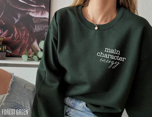 woman wearing forest green sweatshirt with main character energy text seated beside bookshelf and floral artwork