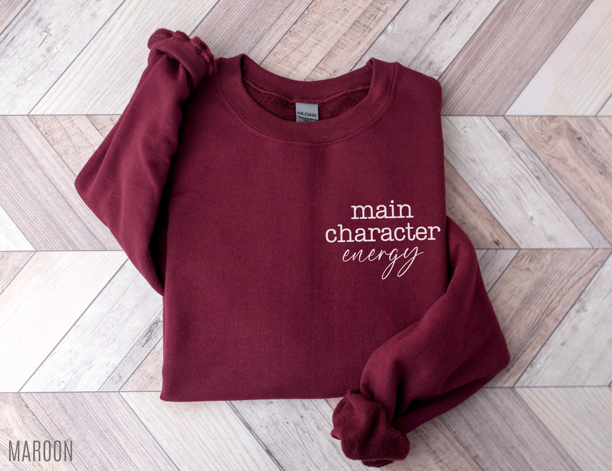 folded maroon sweatshirt with main character energy text on wood herringbone background